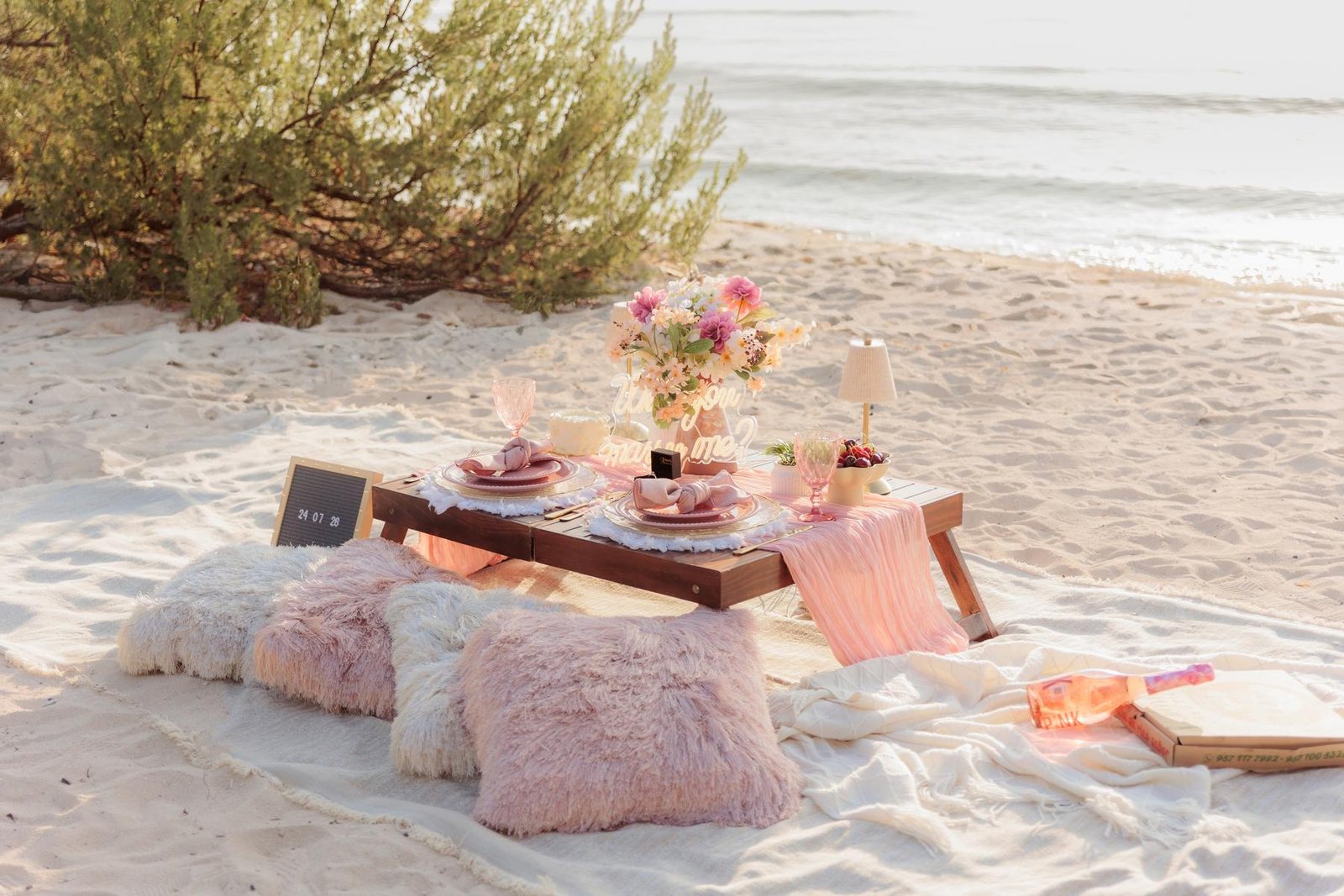 Setup with a “Will you marry me?” sign, florals and pink tableware on the beach